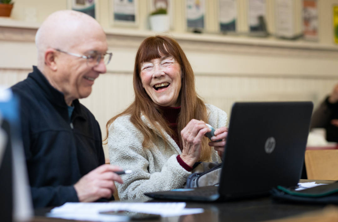 Ein älterer Mann und eine Frau sitzen zusammen an einem Tisch, lächeln und lachen, während sie einen Laptop benutzen. Die Frau hält eine Computermaus, und beide scheinen einen freundlichen, entspannten Moment in einem hellen Innenraum zu genießen.