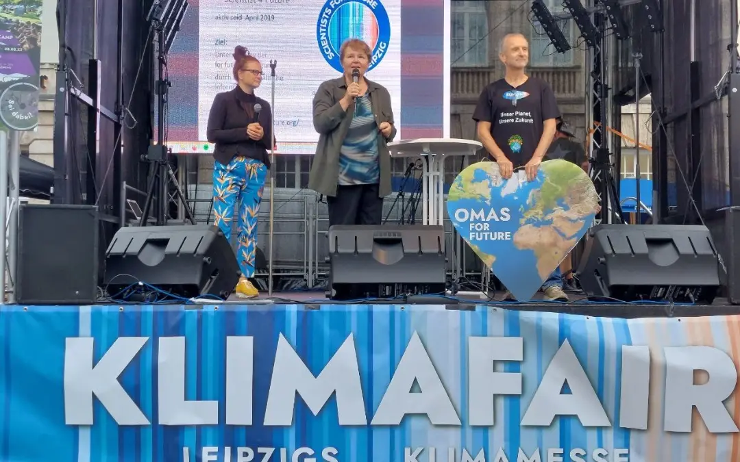 Drei Personen stehen auf einer Bühne bei der KlimaFAIR in Leipzig. Einer spricht in ein Mikrofon, ein anderer hält ein großes herzförmiges Schild mit einer Weltkugel und der Aufschrift OMAS FOR FUTURE. Im Hintergrund ist eine Leinwand zu sehen.