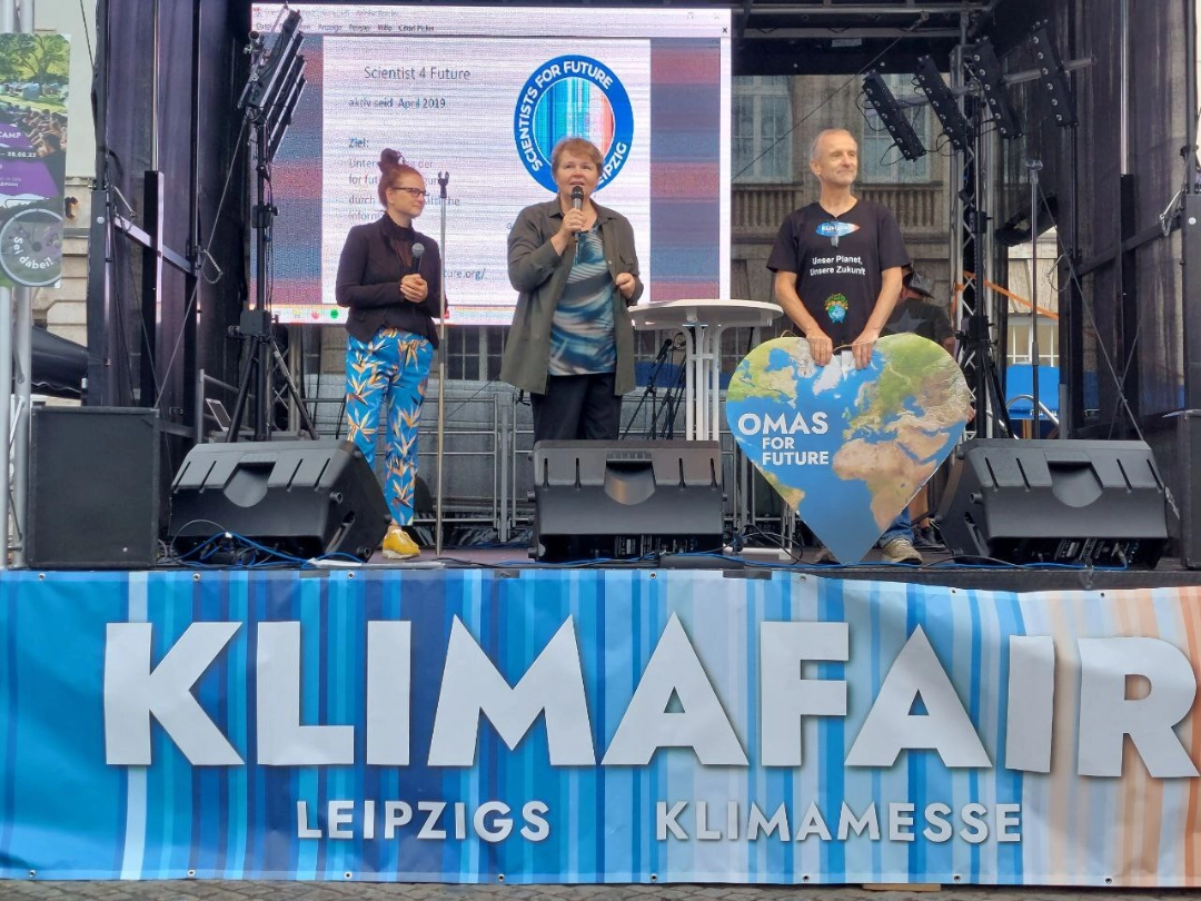 Drei Personen stehen auf einer Bühne bei der KlimaFAIR in Leipzig. Einer spricht in ein Mikrofon, ein anderer hält ein großes herzförmiges Schild mit einer Weltkugel und der Aufschrift OMAS FOR FUTURE. Im Hintergrund ist eine Leinwand zu sehen.