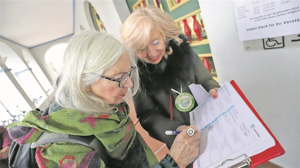 Zwei ältere Frauen stehen in einem Haus; eine hält ein Klemmbrett in der Hand, während die andere ein Dokument unterzeichnet. Beide sind warm gekleidet und scheinen auf den Papierkram konzentriert zu sein. An der Wand neben ihnen ist ein Schild mit Text zu sehen.