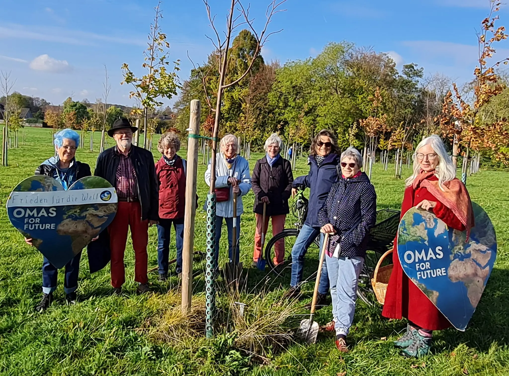 9 Menschen im Freien vor einem neu gepflanztem Baum. Zwei Menschen haben jeweils ein großes Herz, mit der Aufschrift „Omas For Future“, in der Hand.