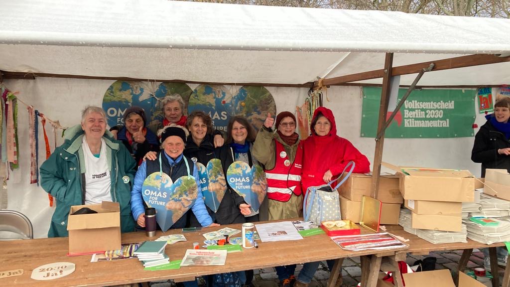 Eine Gruppe lächelnder Menschen steht hinter einem Stand mit Büchern, Flyern und herzförmigen Schildern mit der Aufschrift OMAS FÜR DIE ZUKUNFT. Sie tragen Jacken und Schals und posieren unter einem Vordach bei einer Klimaveranstaltung im Freien.