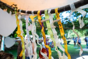 Bunte Bänder mit handgeschriebenen Botschaften in deutscher Sprache sind an einer Stange im Freien befestigt, wobei Menschen und Bäume im Hintergrund verschwimmen und ein Fest oder eine Veranstaltung andeuten.