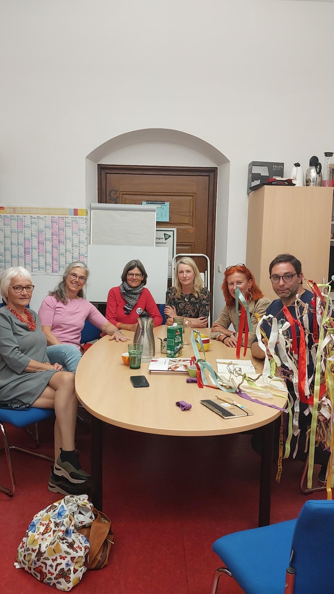 Sechs Erwachsene sitzen um einen runden Tisch in einem Besprechungsraum und lächeln in die Kamera. Verschiedene bunte Bänder, Flaschen, Snacks und Papiere liegen auf dem Tisch. Im Hintergrund sind ein Whiteboard und ein Kalender zu sehen.