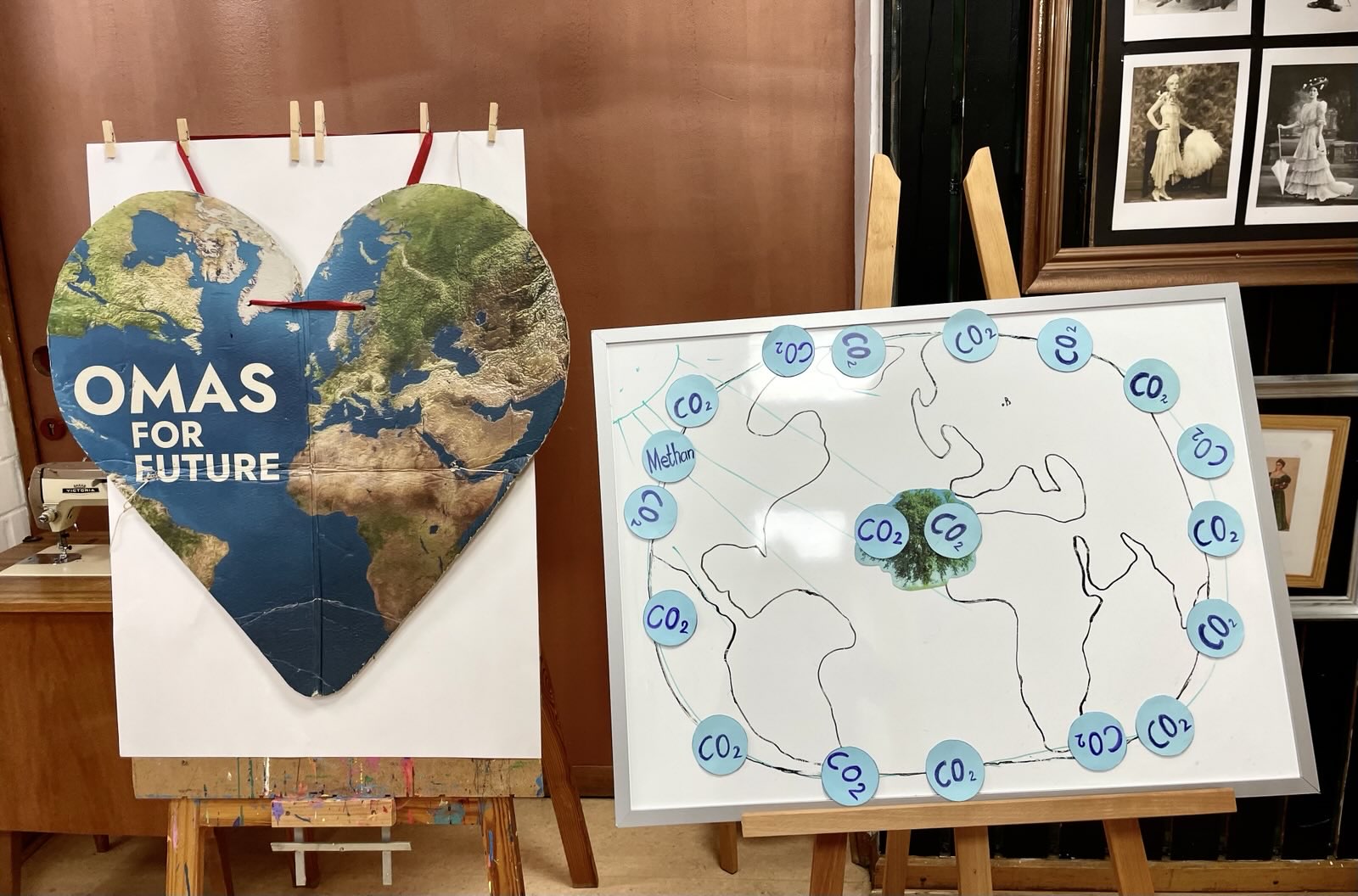 Eine herzförmige Erdkarte mit der Aufschrift OMAS FOR FUTURE steht auf einer Staffelei neben einer Tafelbildzeichnung des Globus, umgeben von blauen CO₂- und Methankreisen, die Themen des Klimawandels hervorheben.
