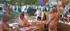 Bei einer Veranstaltung im Freien mit Informationstischen unter schattigen Bäumen kommen die Menschen ins Gespräch. Es werden Banner, Poster und Materialien ausgestellt. Einige Besucher unterhalten sich, während andere sich die Exponate ansehen, wodurch eine lebhafte und anregende Atmosphäre entsteht.