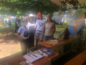 Vier Frauen stehen und lächeln an einem Außenstand von Omas for Future in Bad Kreuznach. Der Stand bietet herzförmige Schilder, Broschüren, umweltfreundliche Artikel und Snacks im Schatten großer Bäume in einer Parkanlage an.