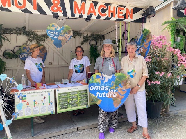 Vier ältere Frauen stehen an einem Klimabewusstseinsstand in Bad Kreuznach, geschmückt mit Pflanzen, Plakaten und einem herzförmigen Omas for Future-Schild. Zwei posieren vorne, zwei stehen hinter einem Tisch mit Infomaterialien.