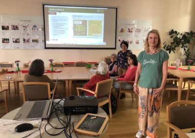 Eine Gruppe von Menschen sitzt an Tischen in einem gut beleuchteten Raum in Bad Kreuznach und sieht sich eine auf eine Leinwand projizierte Präsentation an. Eine Frau steht lächelnd im Vordergrund und trägt ein grünes Smile-Shirt. Auf einem Tisch in der Nähe ist ein Projektor aufgebaut.