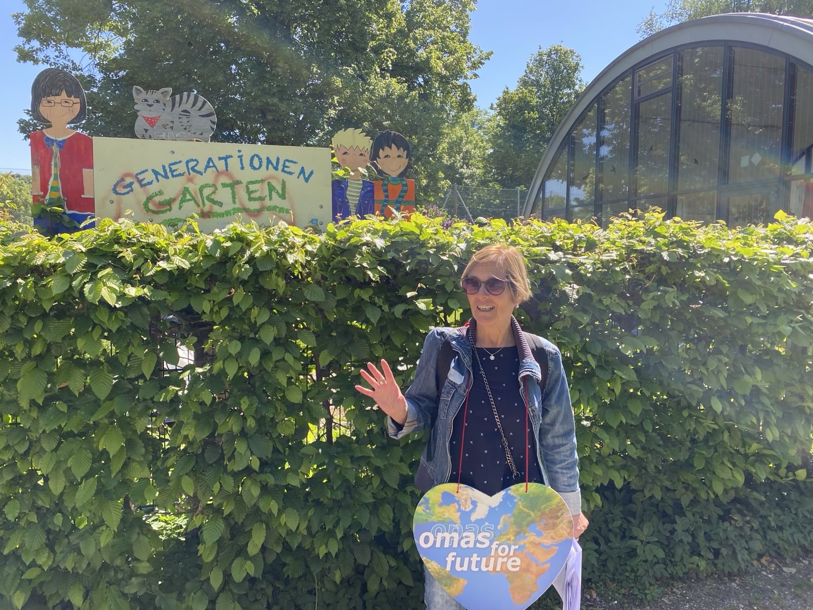Eine Frau winkt und lächelt vor einer Hecke, in der Hand ein herzförmiges Omas for Future-Schild in Bad Kreuznach. Hinter ihr ist ein buntes Generationen Garten-Schild mit Cartoon-Zeichnungen von Menschen und einer Katze zu sehen, sowie Bäume und ein geschwungenes Gebäude.