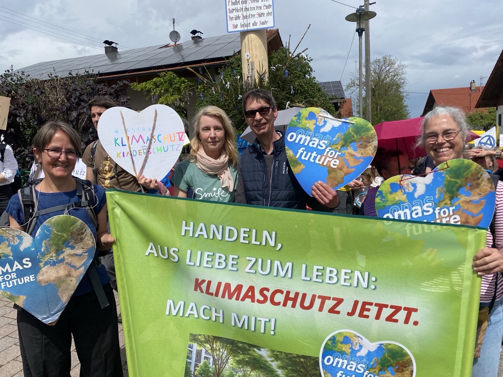 Eine Gruppe von Menschen bei einer Klimaprotestaktion in Bad Kreuznach hält Schilder und ein großes grünes Transparent mit der Aufschrift "Handeln, aus Liebe zum Leben: Klimaschutz jetzt. Mach mit!" Einige halten herzförmige "Omas for Future"-Schilder. Sie lächeln und stehen im Freien.