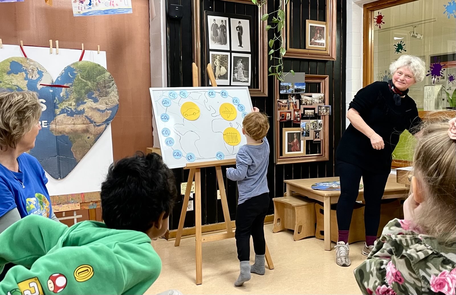 Ein kleines Kind malt auf einer Tafel mit gelben Smiley-Gesichtern, während eine erwachsene Frau in der Nähe lächelt. Andere Kinder und ein weiterer Erwachsener schauen in einem mit Weltkarten und Fotos dekorierten Klassenzimmer zu.