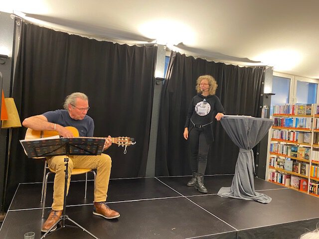 Ein Mann sitzt auf einer kleinen Bühne und spielt Gitarre, daneben steht eine Frau an einem hohen runden Tisch. Hinter ihnen befinden sich schwarze Vorhänge, und auf der rechten Seite sind Regale mit Büchern zu sehen.