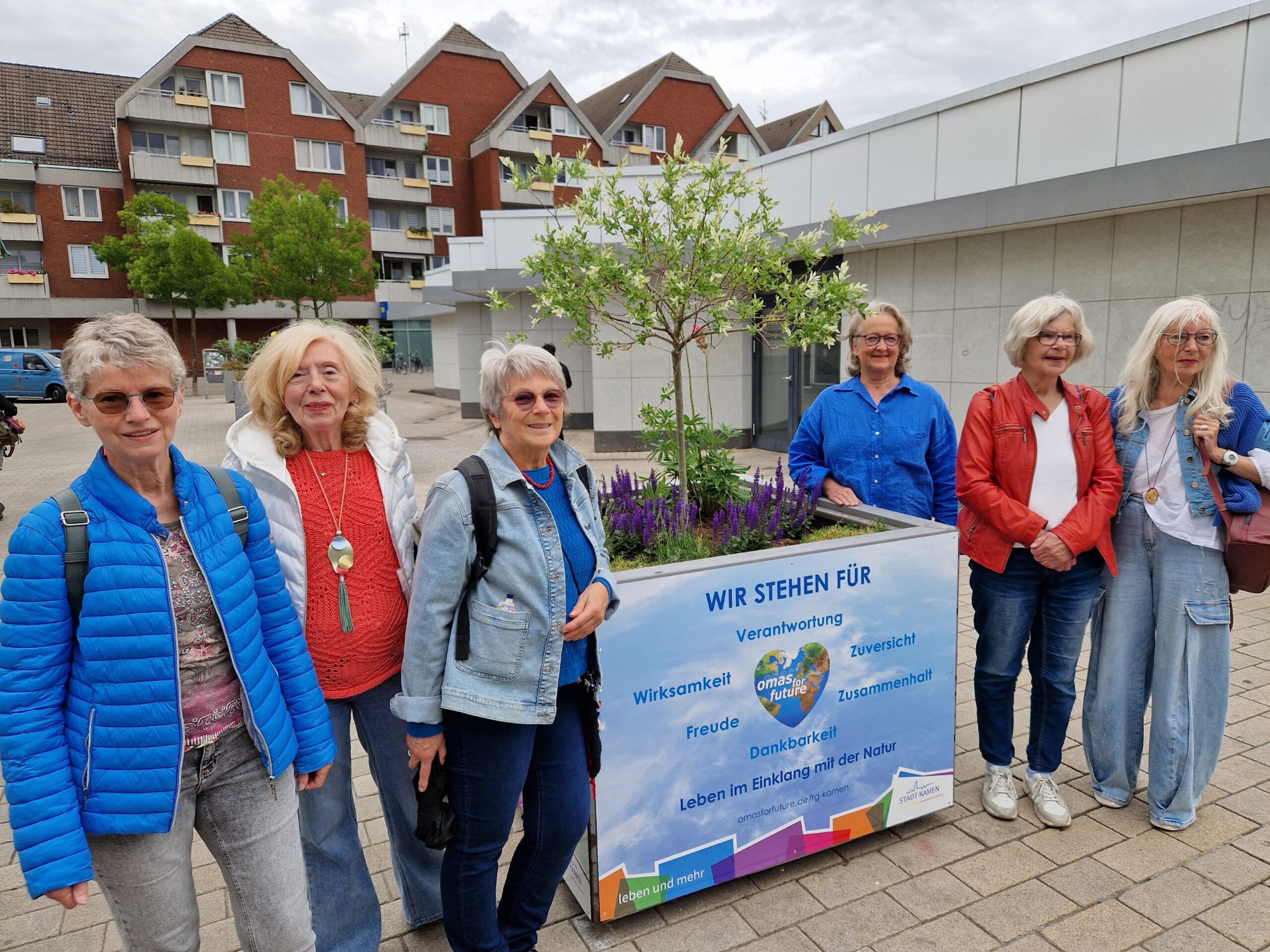 Sechs lächelnde ältere Frauen stehen im Freien auf einer gepflasterten Fläche neben einem Blumenkasten mit einem Schild in deutscher Sprache. Hinter ihnen stehen moderne Gebäude und Bäume. Die Frauen tragen legere Jacken und Brillen.