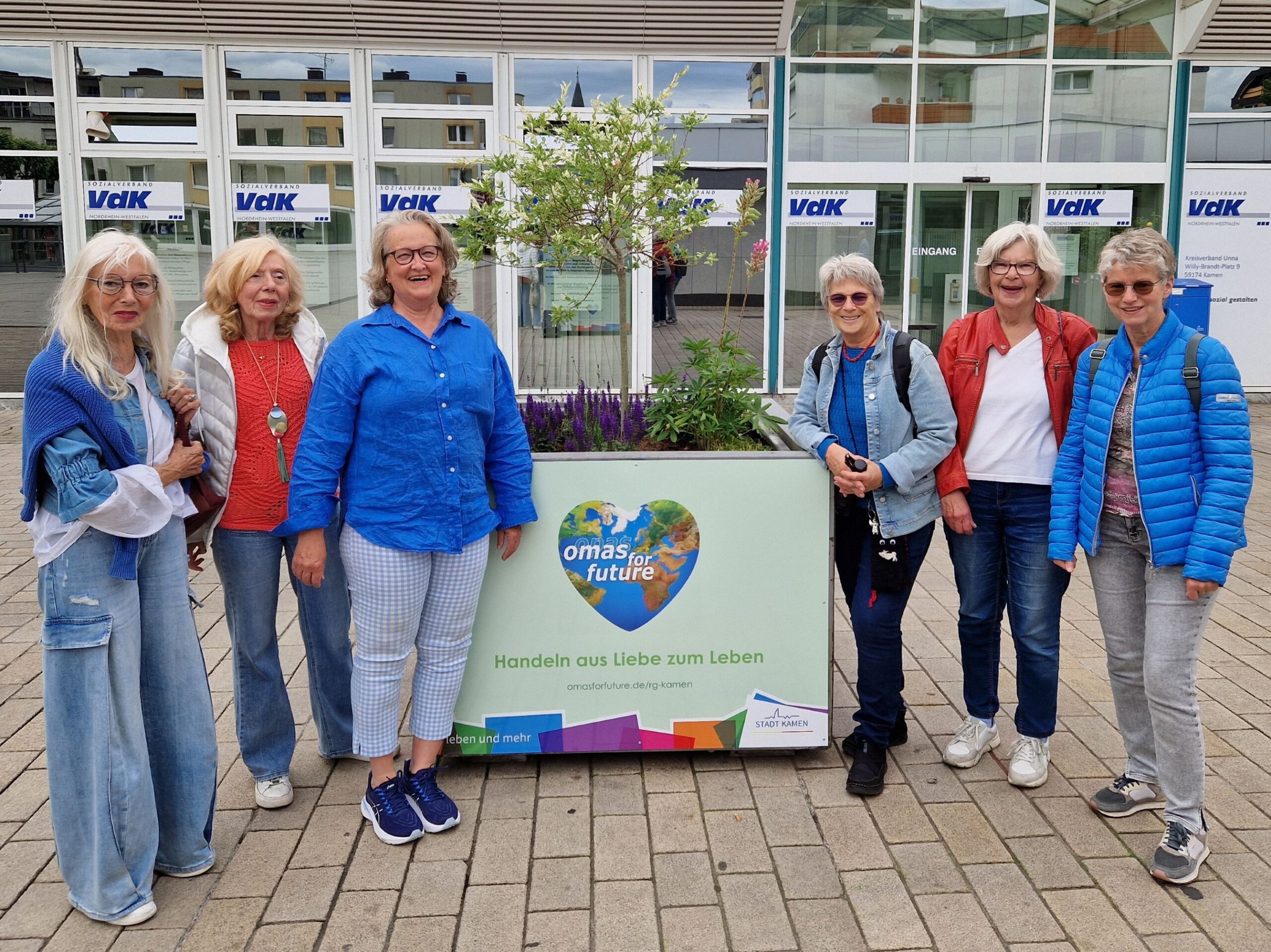 Sechs ältere Frauen stehen lächelnd vor einem Pflanzkübel mit Blumen und einem Schild mit der Aufschrift "omas for future" vor einem modernen Glasgebäude. Sie sind leger gekleidet und wirken fröhlich.
