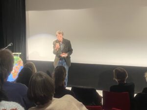 Ein Mann steht auf der Bühne und spricht zu einem Publikum in einem schwach beleuchteten Theater. Er hält eine Notizkarte in der Hand und gestikuliert mit seiner Hand. Die Zuschauer sitzen auf roten Stühlen und schauen ihm zu. Zu seiner Linken befinden sich ein Mikrofon und ein Podium mit einer Weltkugel.