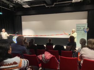 Zwei Personen stehen auf der Bühne vor einem Publikum und halten jeweils ein Ende eines langen, bunten Bandes. Auf der rechten Seite der Bühne ist ein Transparent der Münchner Stadtbibliothek zu sehen.