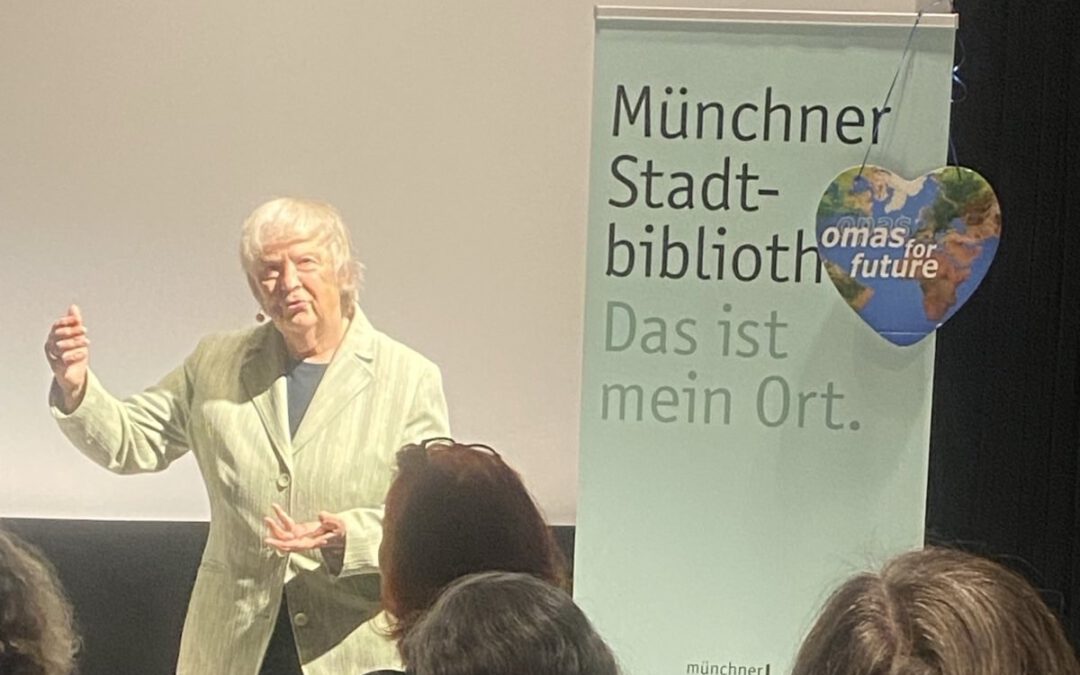Eine ältere Frau spricht vor einem Publikum in der Nähe eines Banners mit der Aufschrift Münchner Stadtbibliothek. Das ist mein Ort. mit einem herzförmigen Schild für Omas for Future daran.