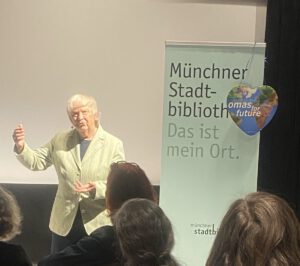 Eine ältere Frau spricht vor einem Publikum in der Nähe eines Banners mit der Aufschrift Münchner Stadtbibliothek. Das ist mein Ort. mit einem herzförmigen Schild für Omas for Future daran.