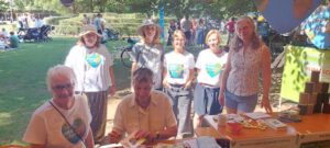 Sieben Erwachsene versammeln sich an einem Tisch im Freien bei einer Gemeinschaftsveranstaltung in Leipzig. Der Tisch ist mit Papieren und Bastelarbeiten bedeckt; einige tragen weiße Hemden mit einem bunten Erdherz-Logo. Im Hintergrund sind Bäume und andere Teilnehmer zu sehen.