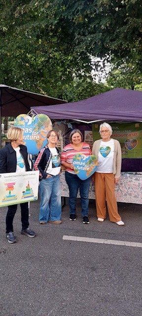 Vier Frauen stehen vor einem Marktstand in Leipzig und halten Umweltschilder und ein herzförmiges "Omas for Future"-Schild. Sie lächeln unter einem Vordach, unter dem im Hintergrund Bäume zu sehen sind.