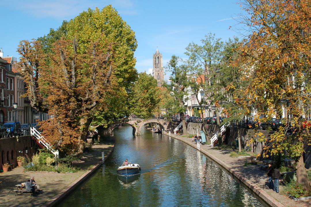 Ein malerischer Kanal, gesäumt von Bäumen und historischen Gebäuden, mit einem kleinen Boot, das auf dem Wasser schwimmt, und Menschen, die am Ufer spazieren gehen. Im Hintergrund erhebt sich ein hoher Kirchturm unter blauem Himmel.
