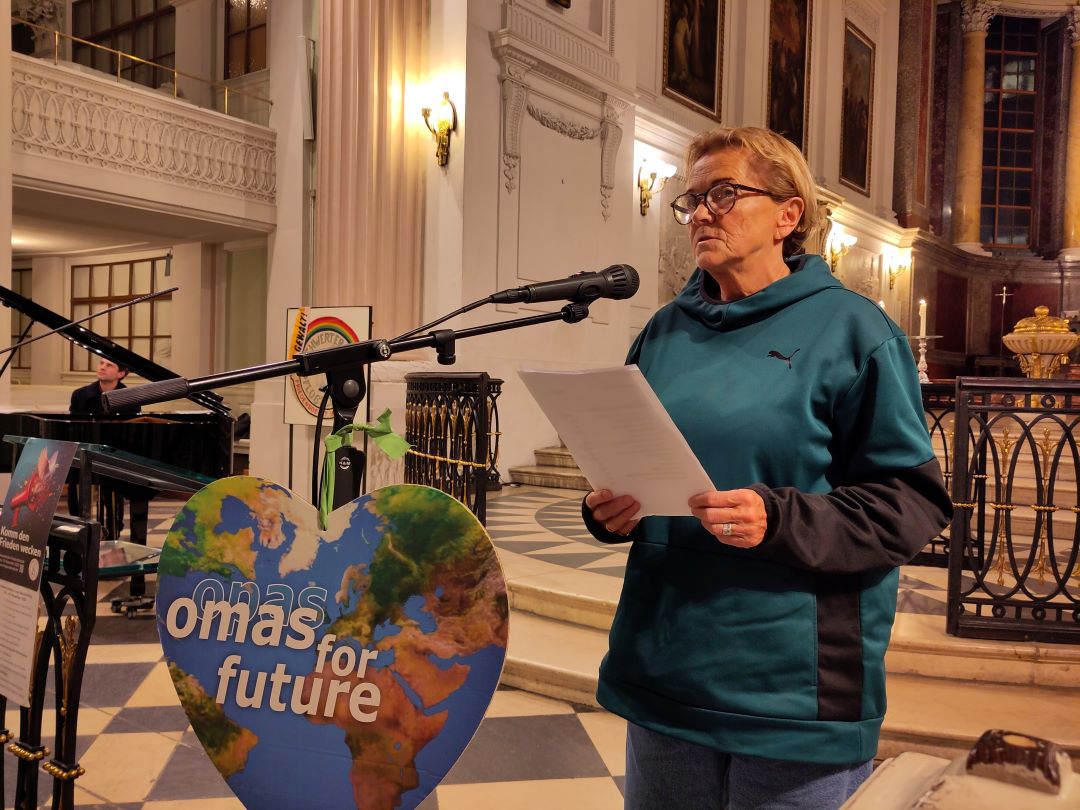 Eine ältere Frau mit Brille und einem blaugrünen Kapuzenpulli steht an einem Mikrofon, hält Papiere in der Hand und spricht in einer Kirche. Vor ihr steht ein herzförmiges Schild mit der Aufschrift omas für Zukunft und einem Bild der Erde.