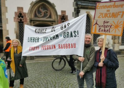 Drei Personen stehen vor einem historischen Gebäude und halten Protestschilder in deutscher Sprache, auf denen sie sich für erneuerbare Energien und gegen die Nutzung fossiler Brennstoffe aussprechen. Eine weitere Frau steht in der Nähe. Im Hintergrund fährt ein Radfahrer vorbei.