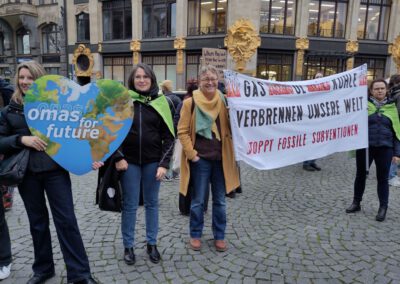 Eine Gruppe von Frauen hält bei einem Klimaprotest ein herzförmiges Omas for Future-Schild und ein Transparent, auf dem die Beendigung der Subventionen für fossile Brennstoffe gefordert wird, während im Hintergrund Gebäude und andere Demonstranten zu sehen sind.