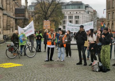 Eine Gruppe von Menschen versammelt sich zu einem Protest auf einem städtischen Platz, einige halten Schilder und Transparente. Einige tragen orangefarbene Sicherheitswesten, und ein Fahrrad steht an der Seite. Im Hintergrund sind Gebäude und Bäume zu sehen.