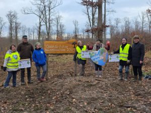 Eine Gruppe von sieben Personen steht im Freien in einem Waldgebiet, einige tragen gelbe Sicherheitswesten. Sie halten Transparente und Schilder mit der Aufschrift "Omas for future" neben einem Holzschild mit der Aufschrift "Omas for future Baumflanzaktion 2025".