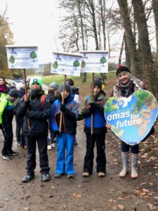 Eine Gruppe von Kindern und ein Erwachsener stehen im Freien und halten Schilder und ein großes herzförmiges "Omas for Future"-Schild, gekleidet für kühles Wetter, mit Bäumen und anderen Menschen im Hintergrund.