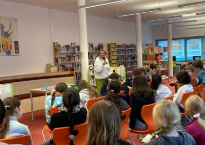 Eine Person steht am Eingang einer Bibliothek und spricht in ein Mikrofon zu einer Gruppe von sitzenden Kindern. Bücherregale säumen die Wände und der Raum ist mit großen Fenstern und orangefarbenen Stühlen hell erleuchtet.