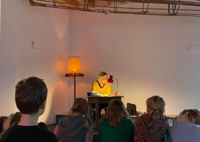 Eine Frau sitzt an einem Schreibtisch unter einer Schreibtischlampe und liest einer Gruppe von Kindern vor, die vor ihr auf dem Boden sitzen. Eine alte Stehlampe leuchtet warm neben ihr in einem ansonsten schummrigen, minimalistischen Raum.