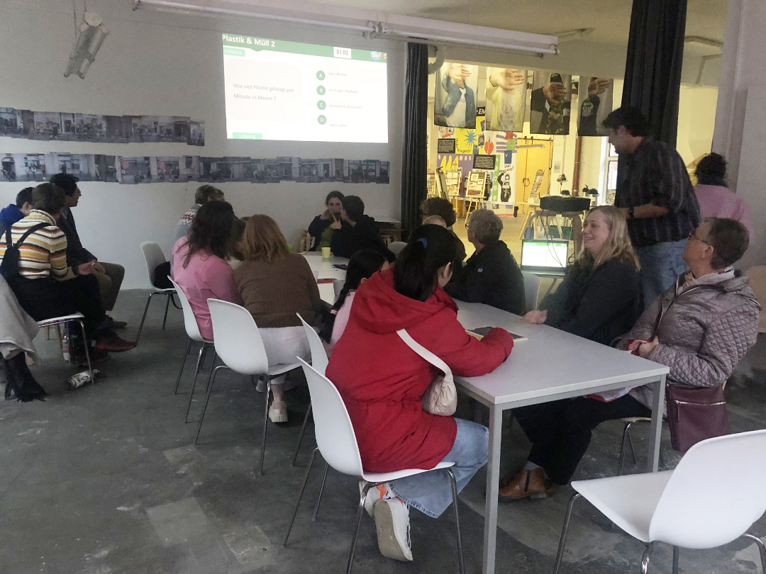 Eine Gruppe von Menschen sitzt an weißen Tischen in einem hell erleuchteten Raum und sieht sich eine Präsentation an, die auf eine Leinwand im vorderen Bereich projiziert wird. Einige Personen sprechen, während andere aufmerksam zuhören.