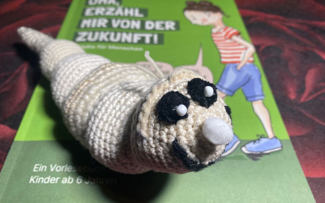 Eine gehäkelte Raupe mit großen Augen liegt auf einem grünen Kinderbuch mit dem Titel "Oma, erzähl mir von der Zukunft!", auf dessen Cover eine Frau und eine Raupe abgebildet sind.