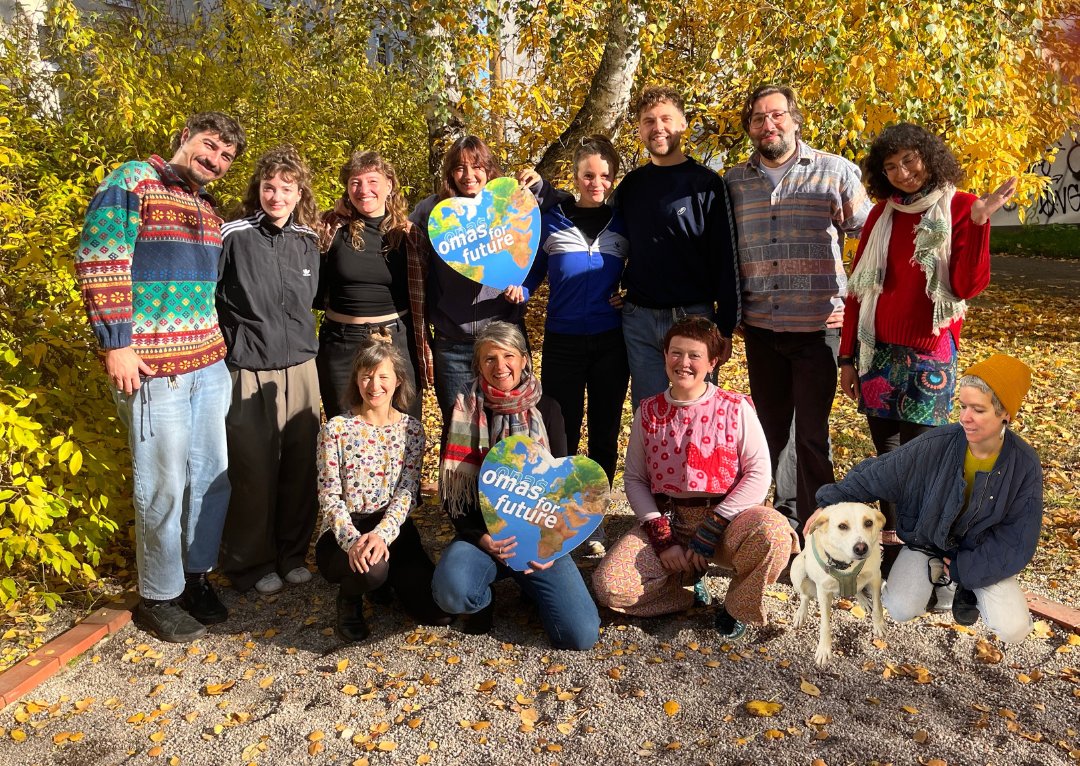 Team Leipzig_omasforfuture Eine Gruppe von dreizehn Personen steht und kniet an einem sonnigen Herbsttag im Freien zusammen und lächelt in die Kamera. Zwei halten herzförmige "Omas for Future"-Schilder in der Hand, und ein gelber Hund sitzt neben ihnen, umgeben von buntem Herbstlaub.