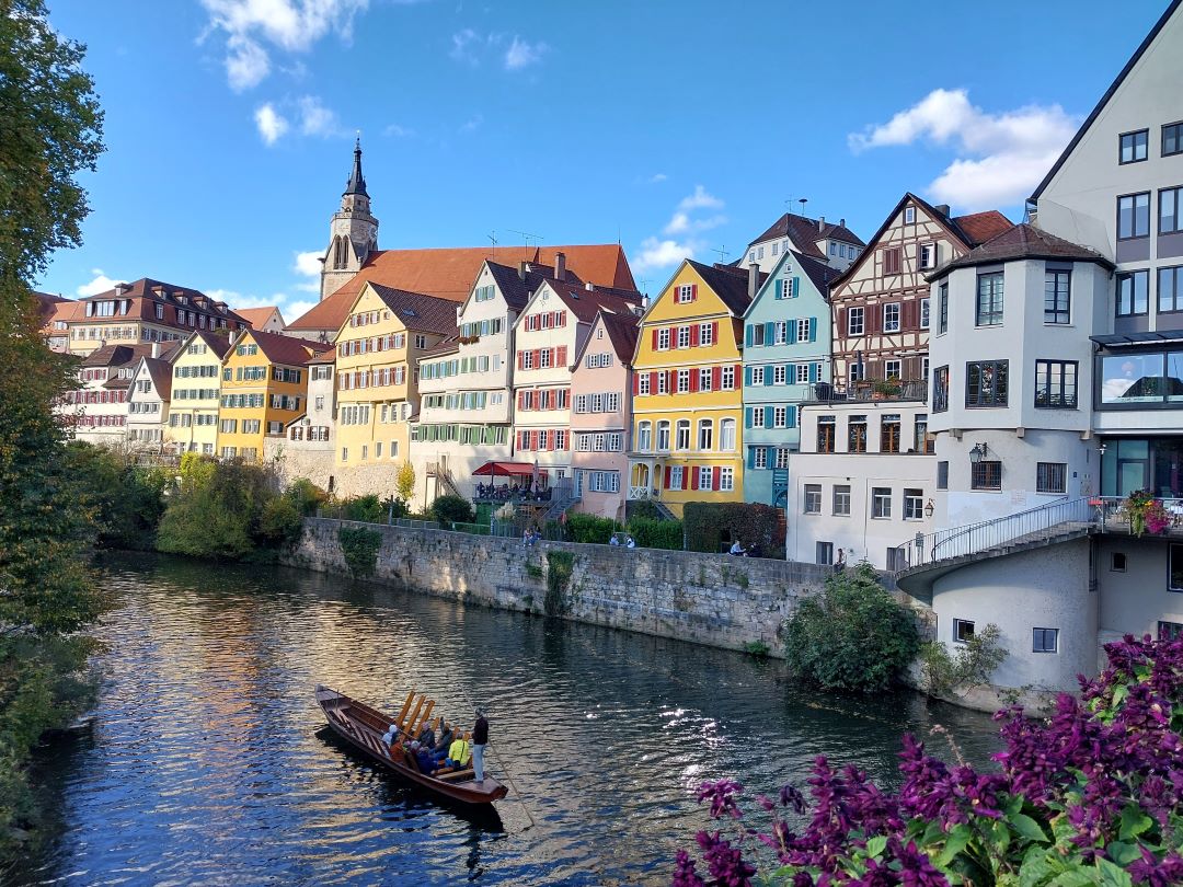Ein kleines Holzboot mit Menschen gleitet auf einem Fluss neben bunten, historischen Gebäuden mit roten Dächern in einer europäischen Stadt, unter einem blauen Himmel mit vereinzelten Wolken. Im Vordergrund sind lila Blumen zu sehen.