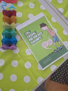 Ein deutsches Büchlein mit dem Titel Oma, erzähl mir von der Zukunft! liegt auf einer grün gepunkteten Tischdecke neben einem bunt bemalten Eierkarton. Das Cover zeigt ein Kind mit einer Lupe, das mit einem Käfer spricht.