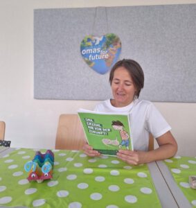 Eine Frau sitzt an einem Tisch mit einer grünen gepunkteten Tischdecke und liest ein Kinderbuch mit dem Titel Oma, erzähl mir von der Zukunft! An der Wand hängt ein herzförmiges Schild mit der Aufschrift Omas für die Zukunft. Auf dem Tisch liegt eine bunte Bastelarbeit.