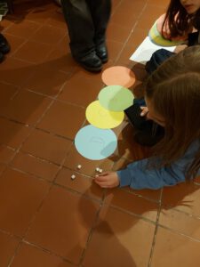 Kinder sitzen und knien auf einem gefliesten Boden und spielen ein Spiel mit farbigen Papierkreisen, die mit Buchstaben beschriftet sind, und kleinen Würfeln. Ein Kind in einem blauen Pullover würfelt in der Nähe der Kreise.