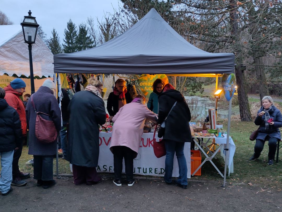 Auf einem Markt im Freien versammelt sich an einem bewölkten Tag eine Gruppe von Menschen um einen Stand unter einem grauen Vordach. Der Stand hat bunte Dekorationen und Produkte, während ein paar Leute in der Nähe sitzen und essen. Im Hintergrund sind Bäume und Gras zu sehen.