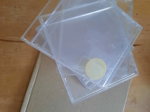 Ein Stapel leerer, durchsichtiger Plastik-CD-Hüllen liegt auf einem geschlossenen Karton, auf einer der Hüllen ist ein kleiner runder Aufkleber zu sehen.