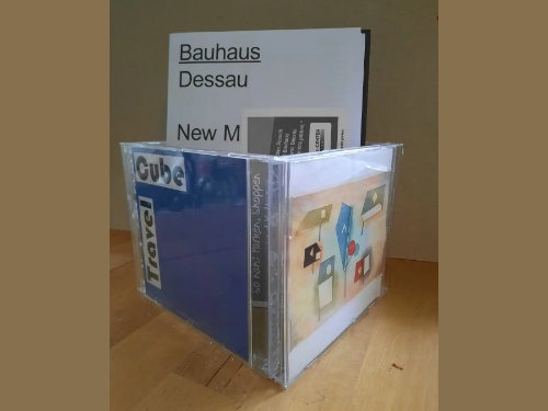 Zwei CD-Hüllen sind auf einer hölzernen Oberfläche abgebildet. Eine CD-Hülle trägt die Aufschrift Cube Travel mit blauem Rücken, die andere zeigt bunte Koffer-Illustrationen. Ein Booklet mit dem Titel Bauhaus Dessau New M ist dahinter teilweise sichtbar.