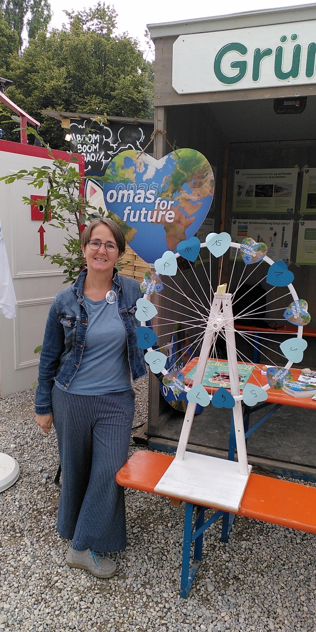 Eine Frau steht lächelnd neben einem dekorierten Spinnrad mit blauen Karten an einem Omas for Future-Stand im Freien in Leipzig. Im Hintergrund sind ein herzförmiges Schild und Grünpflanzen zu sehen.