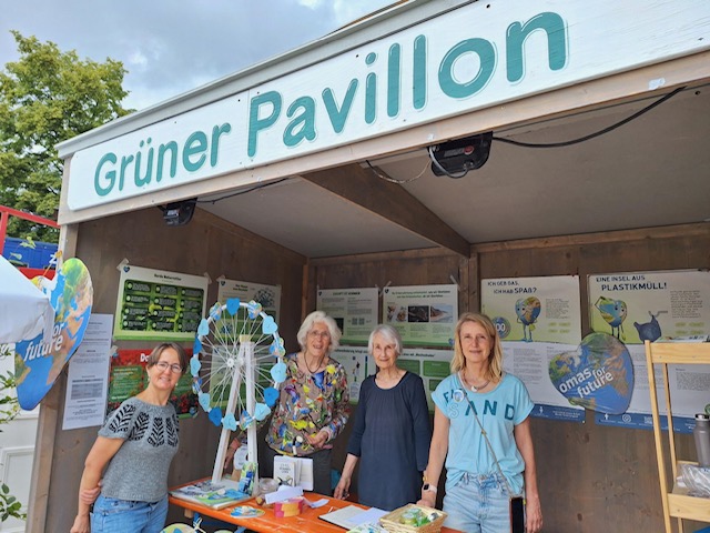 Vier Frauen stehen und lächeln an einem Stand mit der Aufschrift Grüner Pavillon in Leipzig, der mit umweltfreundlichen Schautafeln, Plakaten über Plastikmüll und einem Glücksrad für Umweltbewusstsein wirbt.