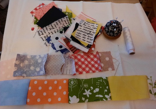 Eine Vielzahl von bunten Stoffquadraten und -rechtecken ist auf einer weißen Fläche angeordnet, zusammen mit einer Spule mit weißem Garn und einem Nadelkissen, das mit Nadeln gefüllt ist. Die Stoffe weisen unterschiedliche Muster und Farben auf.