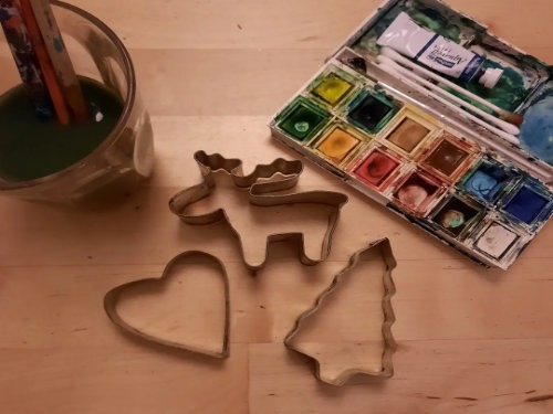 Auf einem Holztisch stehen ein Aquarellfarben-Set mit Pinseln neben einem Glas mit grünem Wasser und drei Ausstechformen in Form eines Herzens, eines Elchs und eines Weihnachtsbaums.