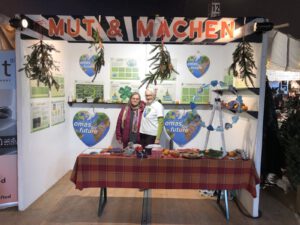 Zwei Frauen stehen lächelnd an einem Stand mit einer rotkarierten Tischdecke, Informationsplakaten und einem Schild mit der Aufschrift MUT & MACHEN. Die Auslage umfasst handgefertigte Produkte, Grünzeug und ein "Omas for Future"-Herzlogo.