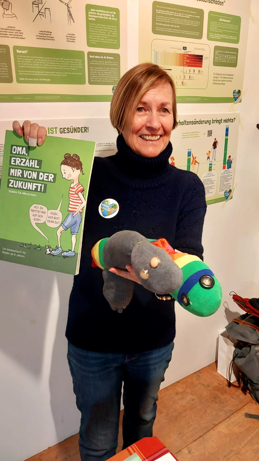 Eine lächelnde Frau, die ein grünes Buch mit dem Titel "Oma, erzähl mir von der Zukunft!" und eine bunte Sockenpuppe in der Hand hält, steht vor Bildungspostern an einer Wand.